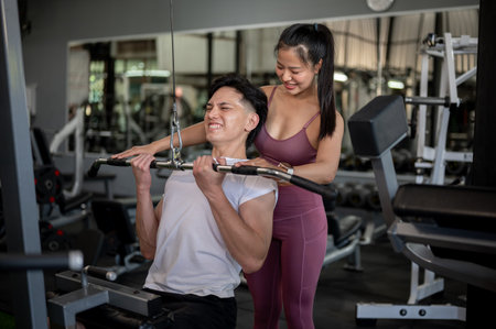 A supportive Asian female fitness trainer is training and instructing a male client in the gym on using the cable machine and maintaining proper form.の写真素材