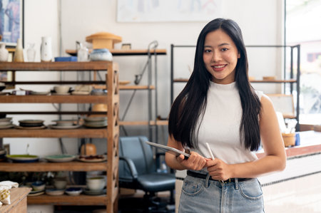 A beautiful Asian female ceramic shop owner or employee stands in her shop with a digital tablet in hand, smiling warmly at the camera.の写真素材
