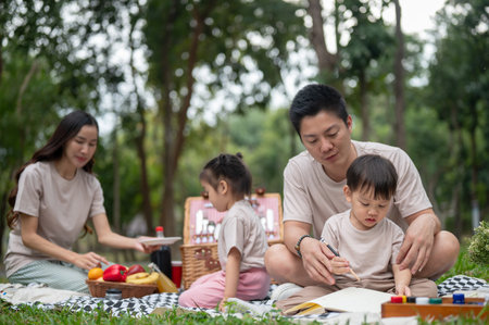 A caring Asian dad teaches his little son to draw and paint on a canvas while enjoying a picnic in the park with their family, as mom and daughter prepare snacks in the background.の写真素材
