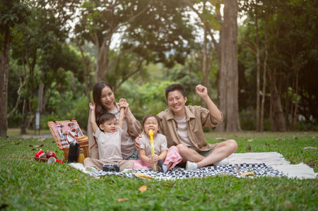 A happy, cheerful Asian family sits on a picnic blanket in a public park, smiling at the camera. fun outdoors time, happy family moment, parenthood, childhoodの写真素材