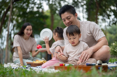 A caring Asian dad teaches his little son to draw and paint on a canvas while enjoying a picnic in the park with their family, as mom and daughter prepare snacks in the background.の写真素材