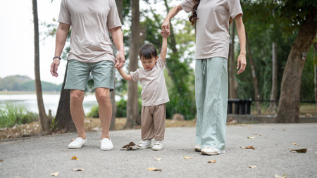 A close-up of a cute, happy little Asian boy holding his parents' hands while walking in a public park, enjoying a morning outdoors. outdoor activity, family moment, childhood, parenthoodの写真素材