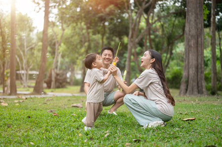 A happy, carefree little Asian boy enjoys playing with bubbles in a green park on a bright day, having a joyful time with his parents. outdoor activity, family moment, childhood, parenthoodの写真素材