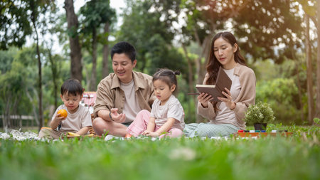 A joyful, happy Asian family picnics in a green public park, sitting on a picnic blanket, enjoying a conversation and eating snacks. happy family, parenthood, preschoolの写真素材
