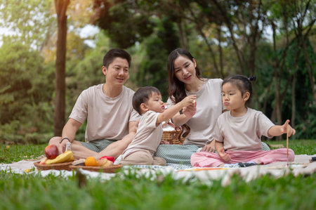 A cheerful Asian family enjoys a picnic in a green public park, sitting on a blanket, eating, and playing with their little boy and girl. happy family moment and outdoors activity conceptsの写真素材
