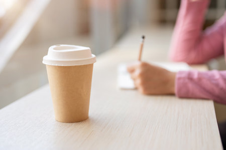 A close-up of a hot drink or coffee cup on a wooden table by a window in a coffee shop, with a blurred background of a woman sitting behind.の写真素材
