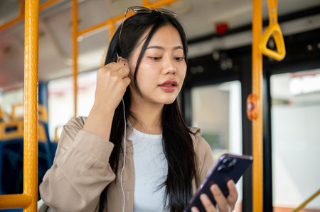 A beautiful Asian woman in casual clothes puts in her earphones, preparing to listen to music during her bus ride. public transportation, lifestyle, city lifeの写真素材
