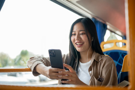 A cheerful, beautiful Asian woman in casual clothes enjoys chatting on her smartphone while sitting at a window seat on a bus, commuting in the city.の写真素材