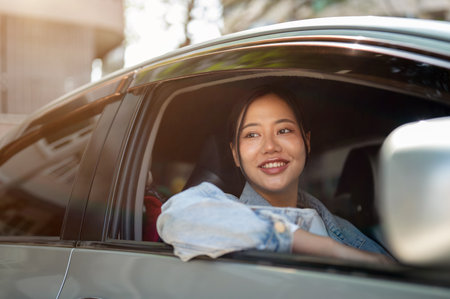An attractive, positive Asian woman in a denim jacket sits in her car, looking out the window and...