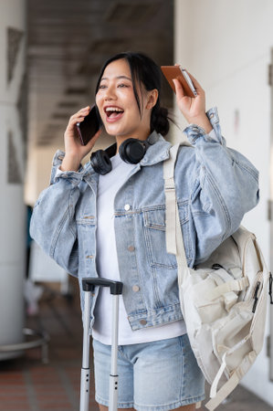 A cheerful, excited Asian female traveler with a backpack and luggage calls her airport taxi while...