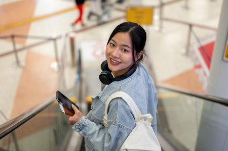 A beautiful, happy Asian female traveler with backpack is on an escalator in the airport terminal,...