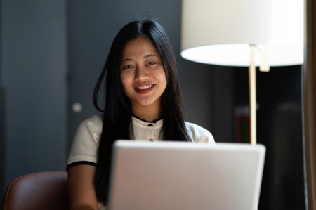 A beautiful Asian woman sits in a coffee shop with her laptop, smiling at the camera. working remotely, e-learning, online meeting, wireless technologyの写真素材