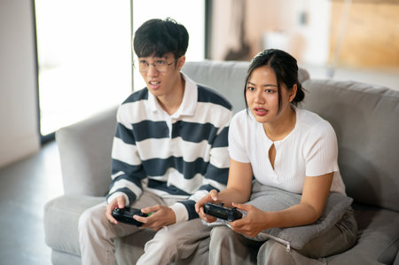 Two young Asian friends, a male and a female, are playing a video game together at home, having a...
