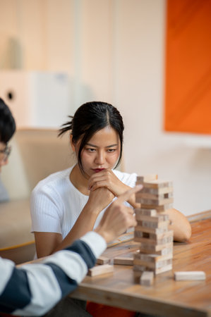 A beautiful, focused Asian woman carefully watches her friend remove a wooden block as they play...