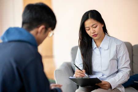 A professional Asian female psychologist or doctor talks to a male patient in her office, listening to his symptoms and noting them in her medical clipboard. mental health, health care, medicalの写真素材