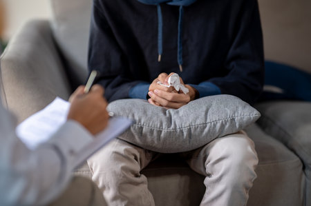 A close-up of a stressed man sitting on a sofa, clasping his hands together in frustration during a mental health checkup with a psychologist. mental health conceptの写真素材