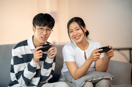 Two excited, joyful young Asian friends are enjoy playing a video game together on the sofa at home, having a fun time. leisure activity, enjoyment, hobbiesの写真素材
