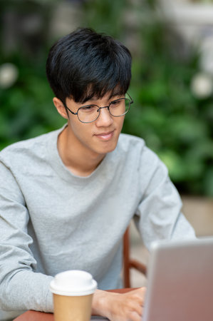 A young Asian man works remotely on his laptop from a coffee shop, sitting at an outdoor table....