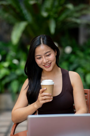 A smiling, beautiful Asian woman enjoys coffee while working remotely on her laptop at a courtyard...