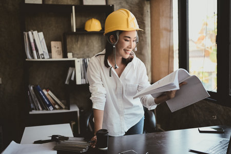 A professional, confident Asian female architect or engineer is reviewing documents at her desk in...