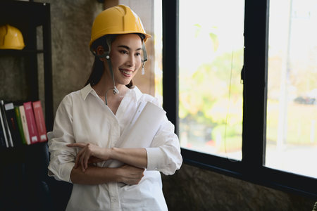 A confident, smiling Asian female architect or engineer boss, wearing a safety helmet, stands in her...