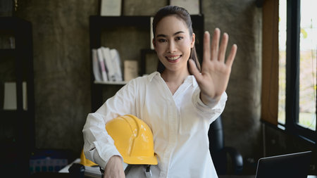 A portrait of a confident, attractive Asian female architect or engineer boss stands in her private office with a safety helmet in her hand, smiling and showing her palm at the camera.の写真素材