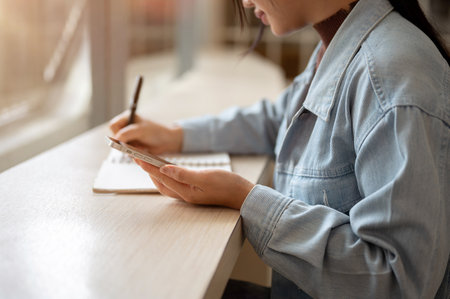 A person holding smartphone while noting down or writing down in the notebook. Taking a note or doing an assignment.の写真素材