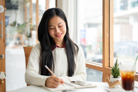 An asian woman is smiling while writing in the notebook, sitting with a tablet and a drink place on the table. study or doing an assignment in the cafe.の写真素材