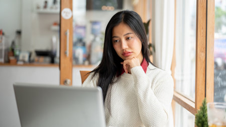 An asian woman is staring intently at the laptop screen, looking focused on work or study. Sitting beside the large wall window of the cafe.の写真素材