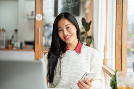 A smiling asian woman is holding the smartphone while looking straight into camera, Sitting with a laptop at the table near a large wall window of the cafe.の写真素材