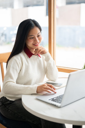An asian woman is smiling while looking at the laptop screen, with one hand under her chin and other holding pencil. Study or working in the cafe.の写真素材