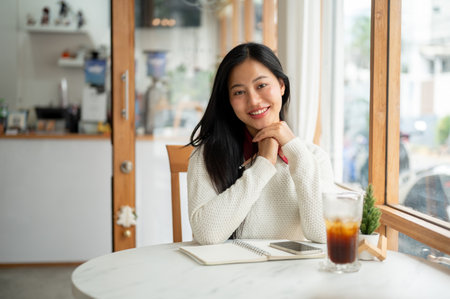 An asian woman is smiling while looking straight into the camera, sitting with a smartphone on notebook and a cold drink at marble table in the cafe.の写真素材