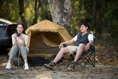 A woman holding a mug and a man are sitting in front of the camping tent. Traveling together, Forest camping, Outdoor activity.の写真素材