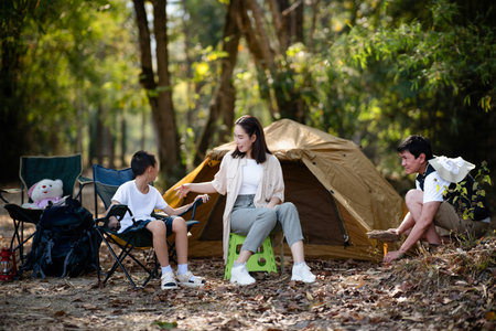 Mother and son is sitting and talking to each other while father setting up their camping tent. Family trip, Forest camping, Outdoor activity.の写真素材