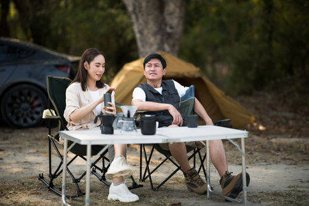 A woman is wiping a water bottle while talking to a man, both sitting at a folding table on a camping ground. They are on a date and are traveling together. Outdoor activity.の写真素材