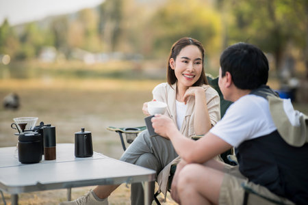 A woman is smiling at the man while holding a cup at folding table, with blurred background of forest scenery. On a date, Travel together, Outdoor activity.の写真素材