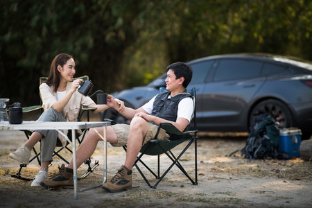 A couple is locking eyes and smiling at each other while pouring the drink at the folding table on the camping ground. Forest camping, Outdoor activity.の写真素材