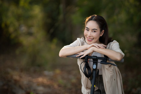 A Smiling asian woman with scooter is looking straight into the camera with the blurred background of forest. Forest camping, Outdoor activity.の写真素材