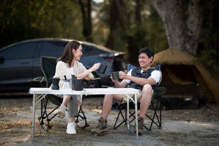 A woman is smiling while pouring drink for the man at folding table in front of their camping tent. Traveling together, Forest camping, Outdoor activity.の写真素材