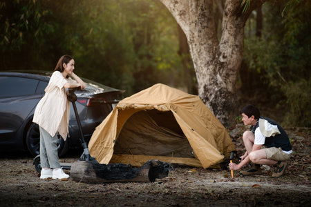 The woman is holding a scooter while smiling to the man setting up their camping tent. Family trip, Forest camping, Outdoor activity.の写真素材