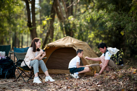 The mother is sitting and watching her son learn how to set up a camping tent from the father. Family trip, Forest camping, Outdoor activity.の写真素材