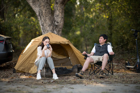 A man and a woman are sitting together in front of the camping tent, The woman is drinking from a mug. Traveling together, Forest camping, Outdoor activity.の写真素材