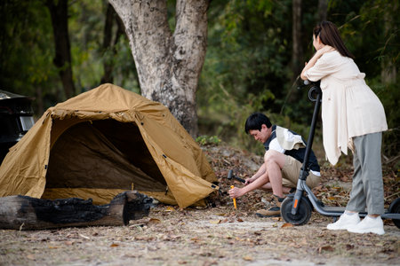 The woman is holding the scooter while watching the man setting up their camping tent. Family trip, Forest camping, Outdoor activity.の写真素材