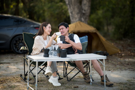 A smiling woman is showing her coffee cup to a man, both sitting at folding table on the camping ground. On a date, Travel together, Outdoor activity.の写真素材