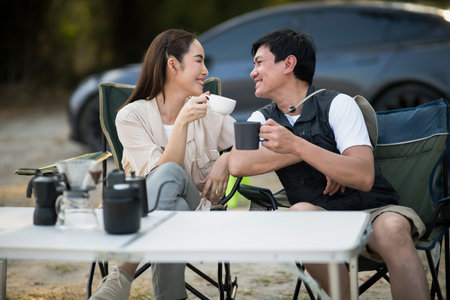 A woman and a man is smiling at each other while holding their cup at folding table on the camping ground. On a date, Travel together, Outdoor activity.の写真素材