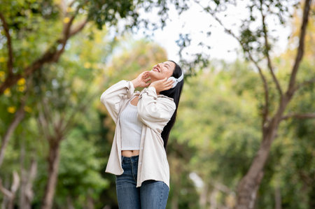 A smiling asian woman is listening to music with headphones while standing in park, breathing in fresh air, Chilling in nature, Outdoor activity, Greenery forest park.の写真素材