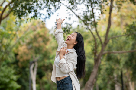 An asian woman is standing with one arm raised while breathing in fresh air. Chilling in nature, Outdoor activity, Greenery park.の写真素材