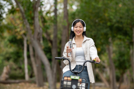 An asian woman is holding a clear water bottle while riding a bike with headphones on. Riding a bike in park, Outdoor activity, Greenery forest park.の写真素材