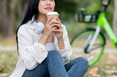 Close up of a asian woman smelling coffee aroma while sitting knees up on park grass field. Chilling in nature, Outdoor activity, Greenery forest park.の写真素材
