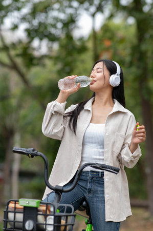 An asian woman is drinking from a clear water bottle while riding a green bicycle with headphones on. Outdoor activity, Greenery forest park.の写真素材